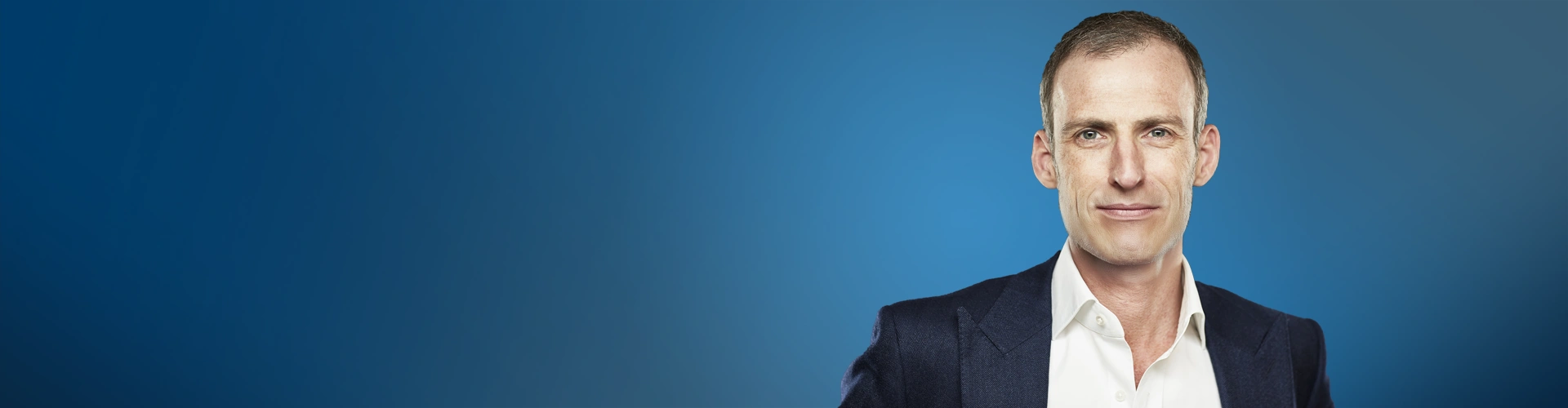 Un primo piano di un uomo d'affari di mezza età con capelli corti e grigi, occhi azzurri e una corporatura snella che fissa dritto con un'espressione neutra. È in piedi, rivolto leggermente verso destra, con il corpo non molto visibile. L'uomo indossa una giacca da abito blu scuro aperta sopra una camicia bianca. Lo sfondo è di un blu profondo sfumato.