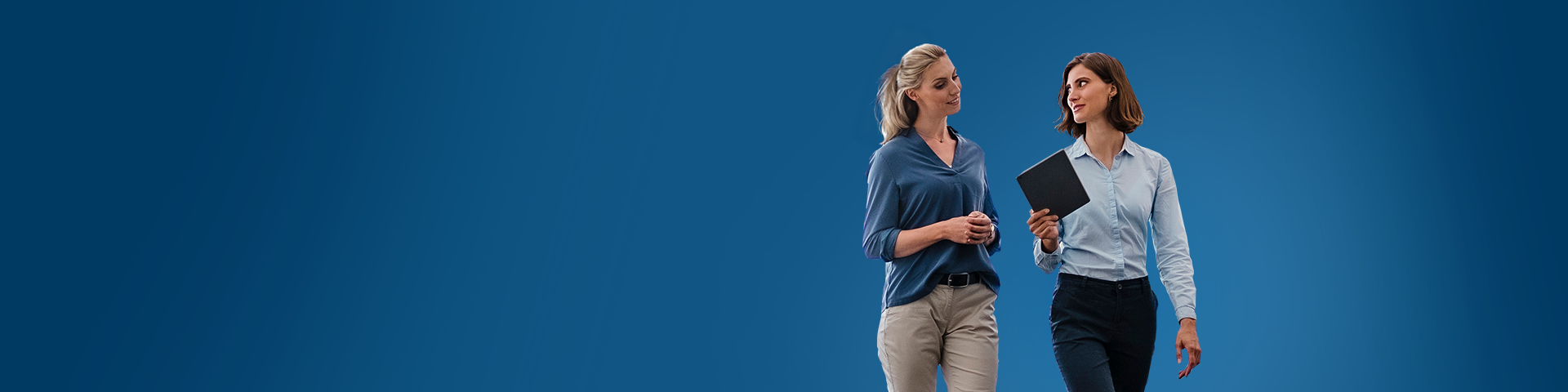 Due donne professioniste a mezzo busto che camminano e sorridono su uno sfondo blu solido. La donna a sinistra ha capelli biondi legati e indossa una camicia di velluto blu, mentre la donna a destra ha capelli castani corti e indossa una camicia azzurra e tiene un tablet nero. Entrambe le donne sono rivolte l'una verso l'altra mentre guardano l'interlocutrice.