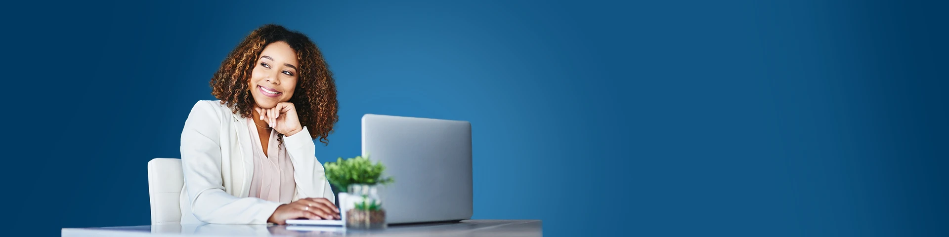 Primo piano di una donna sorridente e con capelli ricci di media lunghezza, vestita con una giacca sartoriale bianca sopra un top color pastello. È seduta a una scrivania di legno chiaro, con la mano appoggiata sulla tastiera di un laptop aperto e il mento appoggiato sull'altra mano. Lo sguardo è rivolto verso l'alto e a sinistra, fuori inquadratura. Sulla scrivania ci sono anche una piccola pianta in vaso e un bicchiere d'acqua. Lo sfondo è un muro blu scuro a tinta unita.