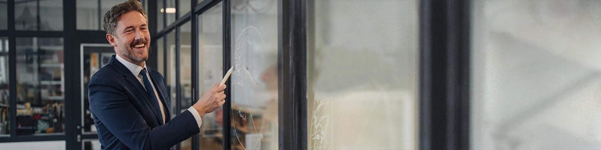 Uomo d'affari sorridente con la barba, che indossa un completo scuro e una cravatta, in piedi in un ufficio moderno con partizioni in vetro. L'uomo è girato verso destra, guardando verso una lavagna trasparente non visibile, e tiene un pennarello bianco nella mano destra come se stesse scrivendo o puntando qualcosa. La sua espressione è gioiosa e coinvolta. L'ambiente dell'ufficio sullo sfondo è leggermente sfocato ma mostra strutture in vetro e metallo.