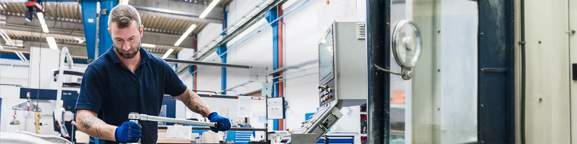 Un uomo con barba e tatuaggio, che indossa una polo blu scuro e guanti da lavoro, opera su un banco da lavoro in metallo. Tiene una lunga leva metallica con entrambe le mani. La scena è ambientata in una moderna officina meccanica con travi metalliche, illuminazione a soffitto, tubazioni e un pannello di controllo della macchina visibile a sinistra.