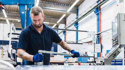 Un uomo con barba e tatuaggio, che indossa una polo blu scuro e guanti da lavoro, opera su un banco da lavoro in metallo. Tiene una lunga leva metallica con entrambe le mani. La scena è ambientata in una moderna officina meccanica con travi metalliche, illuminazione a soffitto, tubazioni e un pannello di controllo della macchina visibile a sinistra.