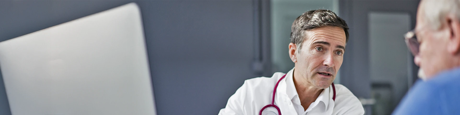 Un primo piano di un medico che parla con un paziente. Il medico indossa un camice bianco e ha uno stetoscopio intorno al collo, mentre il paziente è un anziano sfocato e indossa occhiali.