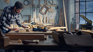 Un artigiano con camicia a scacchi lavora con cura a un banco da falegnameria all'interno di un laboratorio luminoso. Lo sfondo è ricco di strumenti del mestiere, sagome in legno e morsetti appesi alla parete. 