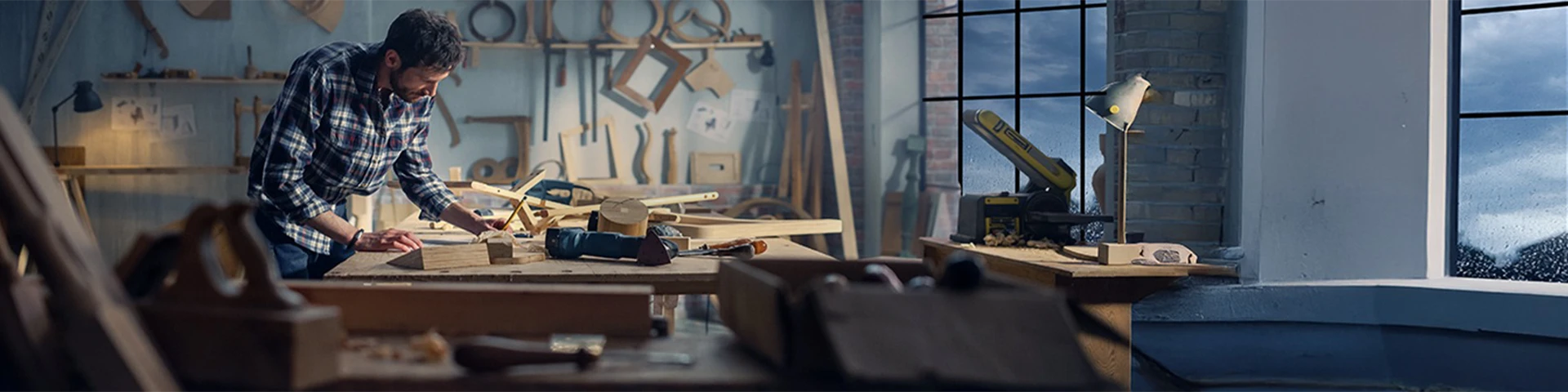 Un artigiano con camicia a scacchi lavora con cura a un banco da falegnameria all'interno di un laboratorio luminoso. Lo sfondo è ricco di strumenti del mestiere, sagome in legno e morsetti appesi alla parete. 