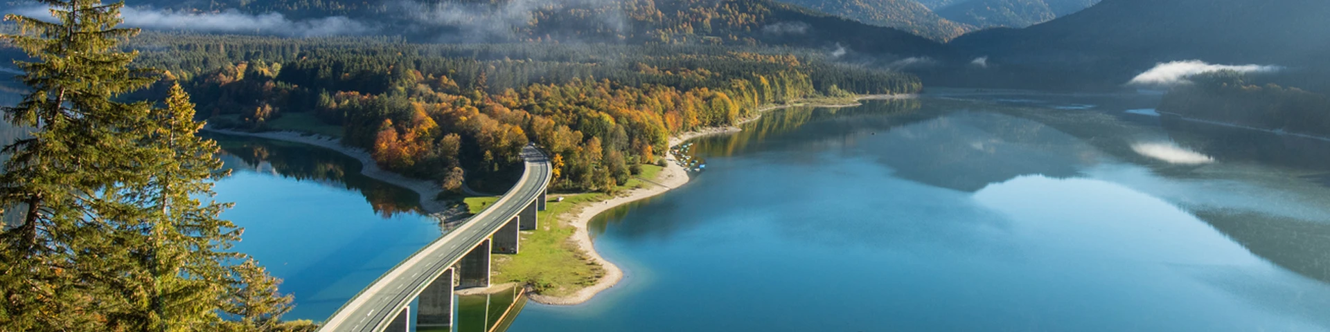 Una veduta panoramica dall'alto di un lago alpino circondato da montagne e boschi con colori autunnali. Un ponte stradale attraversa una parte del lago.