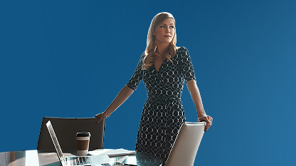 Una donna bionda in abito scuro a fantasia è in piedi dietro una scrivania con un laptop e una tazza di caffè. Guarda verso l'esterno dell'inquadratura con espressione attenta su uno sfondo di un grattacielo per cui alle spalle c’è la vetrata del palazzo.