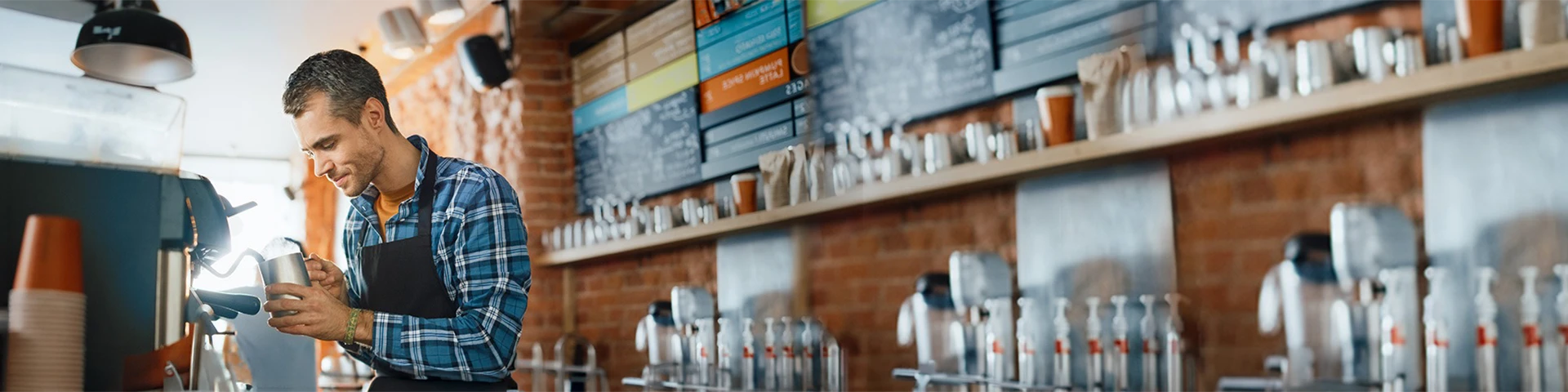 Interno di un bar moderno con pareti in mattoni a vista dove appare un barista. Lui è un uomo di mezza età, con capelli corti grigi e una camicia a quadri blu e bianca sotto un grembiule nero, è ritratto di profilo mentre utilizza una macchina per il caffè espresso professionale. È concentrato nel versare il latte montato da una lattiera in metallo in una tazza, creando una decorazione.
