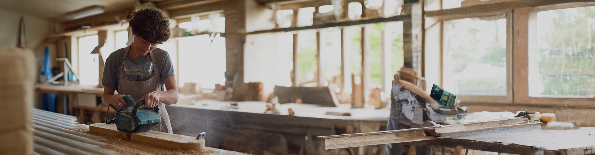 Primo piano di un giovane artigiano al lavoro all'interno di una falegnameria luminosa. Il ragazzo, che indossa occhiali protettivi trasparenti al collo e un grembiule grigio, è concentrato mentre utilizza una levigatrice elettrica su una lunga asse di legno appoggiata su un banco da lavoro. La luce naturale proviene dalle finestre sullo sfondo, piene di attrezzi e segatura.