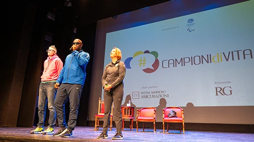 Tre persone su un palco di cui una che parla al microfono e dietro una proiezione con scritto "campioni di vita".